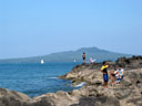 People and fishermen relaxing on the rocks by the sea, Rangitoto Island in background